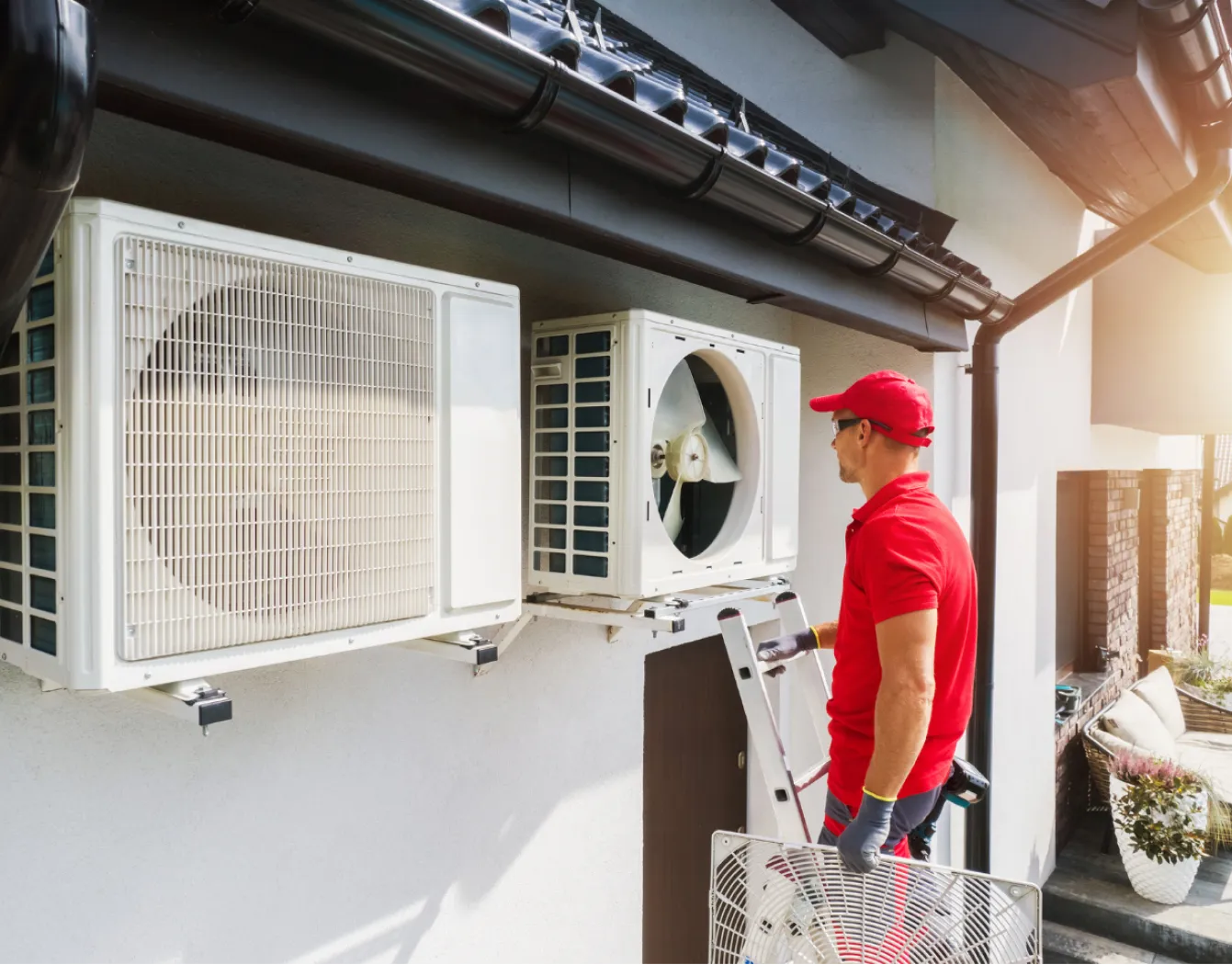 A technician in red uniform services outdoor air conditioning units on a building wall using a ladder.