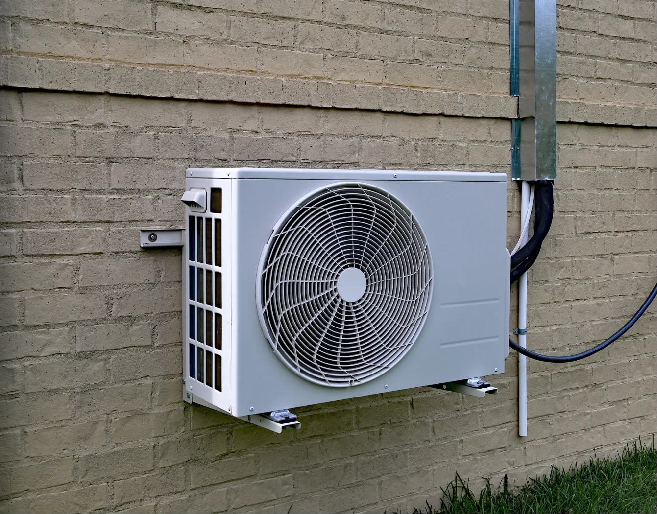 Outdoor air conditioning unit mounted on a brick wall, with electrical conduit attached nearby.