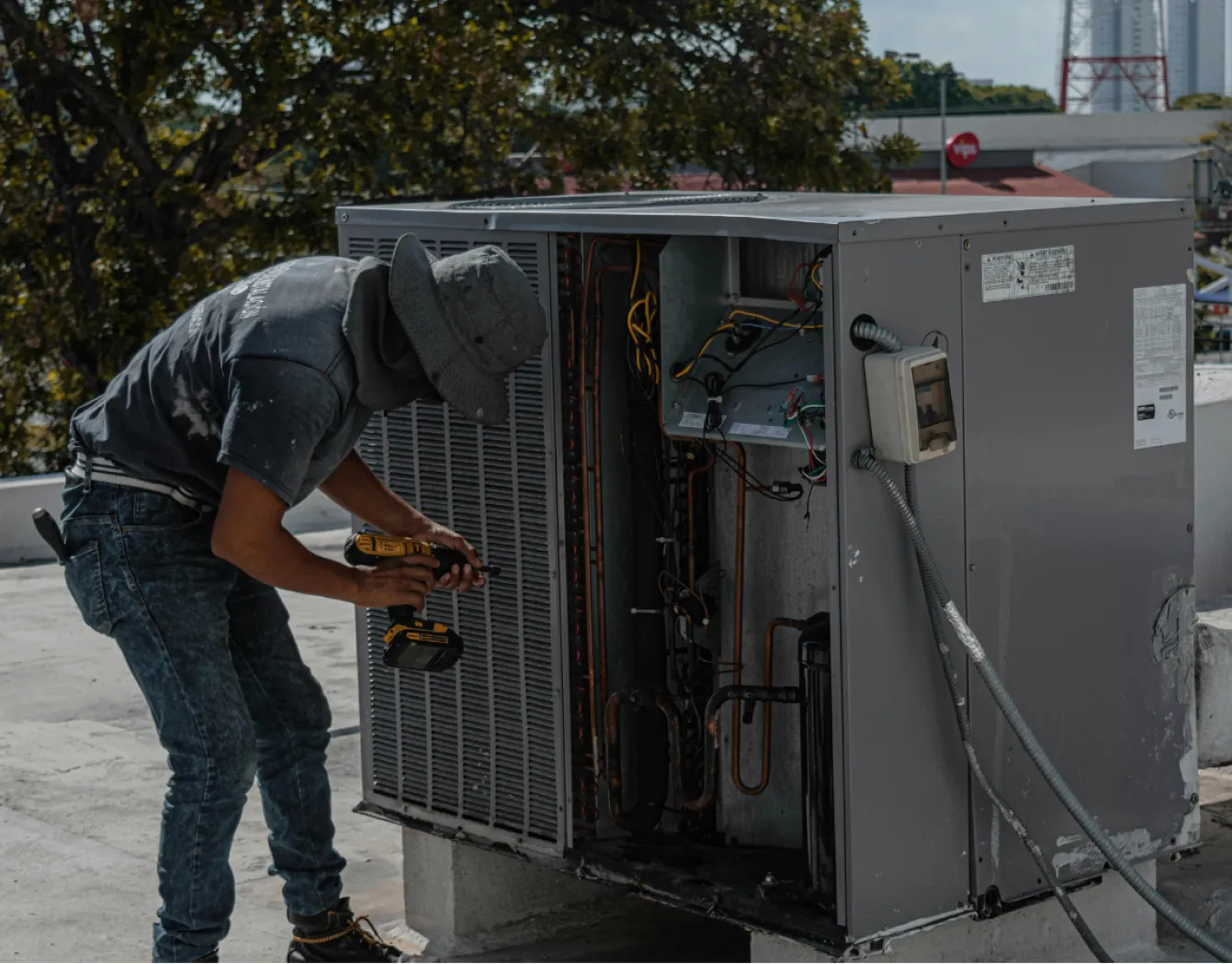 Technician wearing a hat repairs an outdoor air conditioning unit on a rooftop using a power drill.