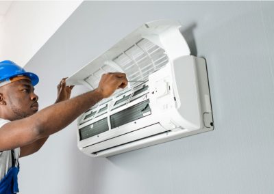 A technician in a blue hard hat repairs a wall-mounted air conditioner with the front panel open.