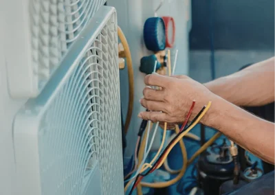 Person repairing an HVAC unit, holding colored wires and tools beside a large outdoor air conditioning unit.