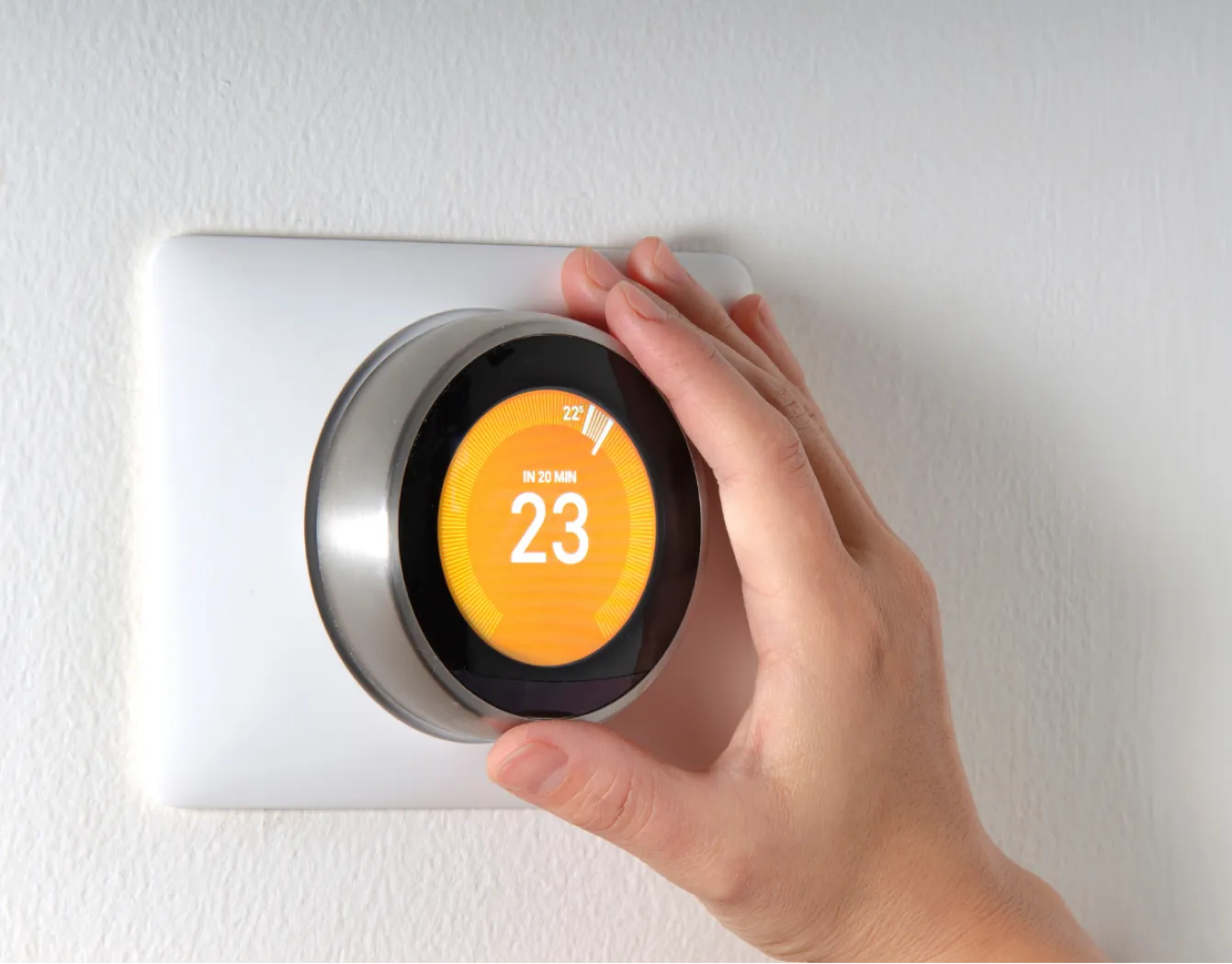 thermostat-1 Hand adjusting a smart thermostat set to 23°C on a white wall.